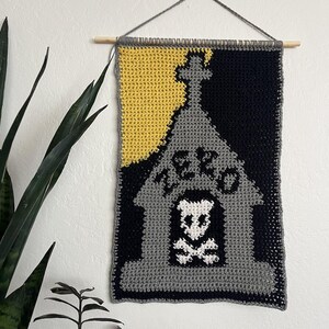 Puede incluir: Tapiz de pared tejido a ganchillo con un diseño de lápida con una calavera y huesos cruzados, el año 1820 y un fondo amarillo y negro. Colgado de una varilla de madera con hilo gris.