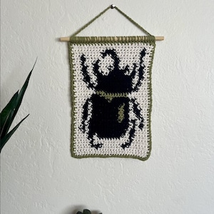 May include: A handmade crochet wall hanging featuring a black scarab beetle design on a white background. The edges are trimmed with green yarn, and it hangs from a wooden dowel with a green yarn loop. A unique piece of home decor.