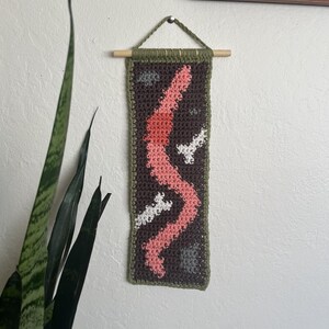 May include: A crocheted wall hanging with a pink, white, and gray abstract design on a brown background. The hanging is framed with a green border and has a wooden dowel at the top.