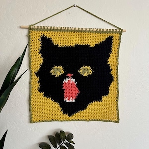 Peut inclure: Une tapisserie murale au crochet représentant un chat noir sur fond jaune. Les yeux du chat sont verts et sa bouche est ouverte, révélant une langue rouge.