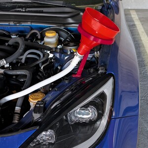 May include: A red funnel with a flexible white tube is positioned over the engine of a blue car. The car's hood is open, revealing the engine components. The car's headlight is visible.
