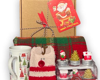 May include: A Christmas gift box containing a red tartan throw blanket, a white mug with a Christmas tree design, a pair of red and white striped reindeer socks, and a set of four small Christmas-themed candles.