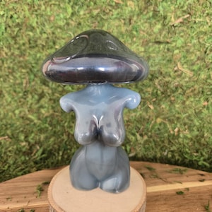 May include: A gray ceramic figurine of a woman with a mushroom-shaped head. The figurine is sitting on a wooden base.