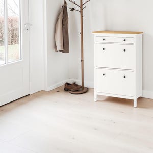 Peut inclure: Un meuble à chaussures blanc avec un dessus en bois clair et des boutons noirs. Un manteau marron est suspendu à un porte-manteau en bois. Une paire de chaussures marron est posée au sol. Le sol est en bois clair. Le meuble mesure environ 80 cm de haut.