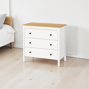 Può includere: Un comò bianco a tre cassetti con piano in legno chiaro e pomelli neri. Il comò è appoggiato a una parete bianca e su un pavimento in legno chiaro. L'arredamento della camera da letto è semplice e moderno.