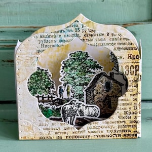 Puede incluir: Una caja de papel decorativa con una escena recortada de un paisaje rural. La caja está hecha de papel envejecido con texto, con una casa, árboles y figuras. La caja tiene una parte superior arqueada y una base rectangular.