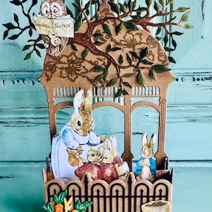 May include: A whimsical papercraft scene featuring Peter Rabbit and family. The scene includes a brown wooden structure with an owl reading a book labeled "Squirrel Nutkin." Rabbits are gathered inside, with a fence and a basket of carrots in the foreground.