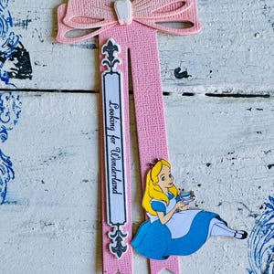Könnte beinhalten: Ein rosa Lesezeichen mit einer Schleife und einem dekorativen Design. Das Lesezeichen zeigt den Text "Looking for Wonderland" und eine Illustration von Alice im Wunderland. Das Lesezeichen ist aus Papier.