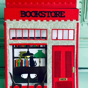 May include: A red and white papercraft bookstore with the word "BOOKSTORE" across the top. The building features a window display with books, a black cat, and a table with two cups. A red door is on the right.