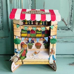 Può includere: Un modello di carta in miniatura di un negozio "Green Grocers" con una tenda a strisce rosse e bianche. Il negozio è decorato con ghirlande colorate, piante in vaso e illustrazioni di personaggi animali. Una scena affascinante e stravagante.