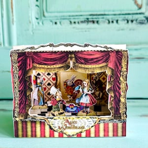 Peut inclure: Un diorama pop-up représentant des scènes d'Alice au pays des merveilles. La scène a des rideaux rouges, une cantonnière dorée et des illustrations de personnages comme Alice, le Chapelier fou et le Chat du Cheshire. La base a des rayures rouges et dorées, avec le texte "Alice in Wonderland".