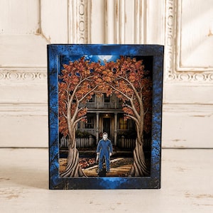 May include: A rectangular shadow box featuring a layered paper diorama of a house at night. Two large trees with orange autumn leaves frame the scene. A figure in a blue jumpsuit stands in the foreground, holding a knife in front of the house.