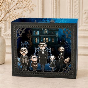Può includere: Una shadow box rettangolare che presenta una scena in carta ritagliata a strati con una famiglia di cinque personaggi davanti a un maniero blu scuro. La cornice è nera con ritagli decorativi a ragnatela ed è visibile una mano.