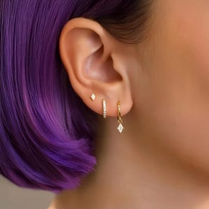 May include: Close-up of an ear adorned with three gold earrings. The top earring is a small diamond shape stud. The middle earring is a small hoop with a row of small diamonds. The bottom earring is a small gold hoop with a diamond-shaped charm.