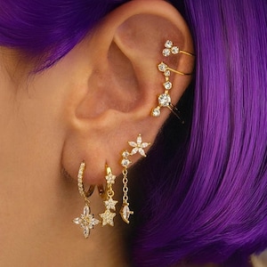 Op de afbeelding: Een verzameling goudkleurige oorbellen en oor manchet. De oorbellen hebben ster- en bloemmotieven met heldere edelstenen. Eén oorring heeft een sterhanger. De oor manchetten hebben een delicaat, gelaagd ontwerp met sprankelende accenten.