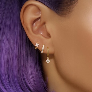 May include: A close-up of an ear adorned with three gold earrings. One earring is a small flower stud, another is a small hoop with a row of clear stones, and the third is a gold hoop with a dangling four-point star design with clear stones.