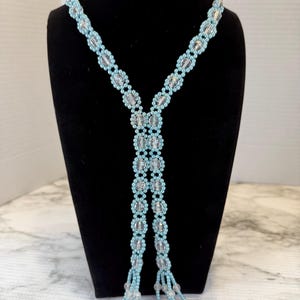 Könnte beinhalten: Eine Lariat-Halskette aus hellblauen, kugelförmigen Perlen und klaren, facettierten Kristallperlen, die in einem sich wiederholenden Blumenmuster angeordnet sind. Das Design besteht aus zwei langen Strängen, die in Perlenquasten enden.