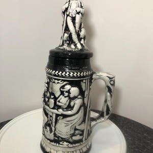 May include: A black and white ceramic beer stein with a lid featuring a figurine of a man in traditional clothing. The stein has a handle and is decorated with a scene of people in a village setting.