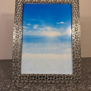 Può includere: Una cornice rettangolare in metallo argentato con un bordo a motivo floreale. La cornice contiene un'immagine di un cielo blu con nuvole bianche e un sole luminoso. La cornice è esposta su una superficie grigia.