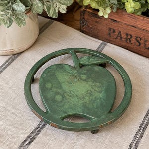 May include: A green cast iron trivet in the shape of an apple. The trivet has a circular frame and a detailed apple design. The trivet is on a striped linen cloth. The trivet is designed to protect surfaces from hot cookware.