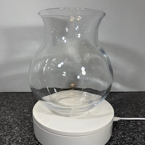 May include: A clear glass vase with a rounded body and flared rim sits on a white, rotating platform. The vase has a wide opening and a smooth, transparent surface, suitable for floral arrangements or decorative displays.