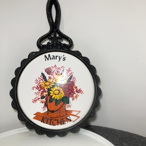 Puede incluir: Salvamanteles de hierro fundido negro con un inserto de cerámica blanca. La baldosa tiene un diseño floral con un cubo de flores y el texto "Mary's Kitchen".