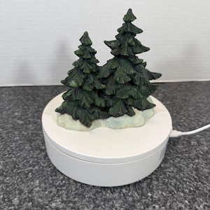 May include: A miniature Christmas scene featuring two dark green fir trees with snow accents, set on a white rotating base. The trees have detailed, textured branches. A white cord extends from the base.