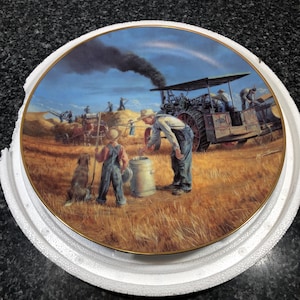 Puede incluir: Plato decorativo con una escena de cosecha rural con un tractor a vapor, agricultores y un perro. El plato tiene un borde dorado y está colocado en un soporte protector blanco. La obra de arte representa un campo de trigo bajo un cielo azul.