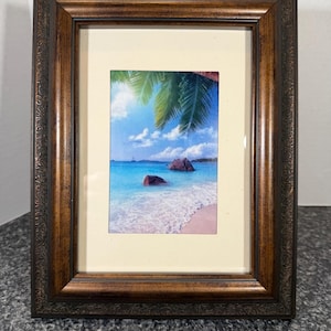 May include: A framed print featuring a tropical beach scene with turquoise water, white sand, and palm trees. The print is set within a dark brown wooden frame with ornate detailing, creating a classic aesthetic.