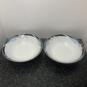 May include: Two white ceramic bowls with a black rim and floral design. The bowls have a black border with a pattern of blue, pink, and green flowers. The bowls are empty and sit on a dark speckled surface.