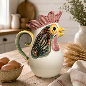 Puede incluir: Jarra de cerámica con forma de gallo, cuerpo blanco, pico amarillo y cresta roja. La jarra tiene un asa verde y amarilla y está decorada con detalles verdes, marrones y rojos. Está sobre una mesa de madera con un cuenco de huevos y una servilleta de lino.