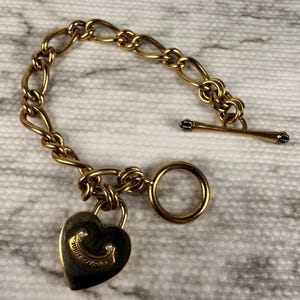 May include: A gold-tone chain link bracelet featuring a toggle clasp and a heart-shaped padlock charm. The padlock is engraved with the text Juicy Couture and includes two small clear stones set into the metal surface of the charm.