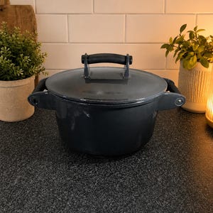May include: A dark gray cast iron Dutch oven with a lid and two side handles. The pot has a black handle on top and sits on a dark speckled countertop. The background includes a white tiled wall and decorative plants.