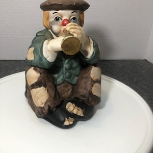 Puede incluir: Una figura de payaso de cerámica en posición sentada, tocando un pequeño instrumento de latón. El payaso lleva un sombrero marrón, una chaqueta verde y pantalones marrones remendados. El payaso tiene una nariz roja y rasgos faciales pintados.