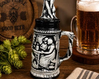 May include: A black and white ceramic beer stein with a detailed relief design. The stein has a handle and a decorative lid with a figurine on top. A pint of beer and a wooden barrel with the text "GOOD PEOPLE GOOD BEER" are in the background.