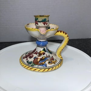 May include: A white ceramic candlestick with a yellow, blue, and red floral design. The candlestick has a handle and a single candle holder.