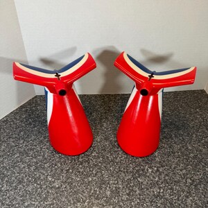 May include: Two identical ship funnel replicas featuring a vibrant red base, a white vertical stripe, and a distinctive blue and white winged top section. Each funnel has a smooth, glossy finish and a circular opening at the center of the top.