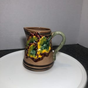May include: A small brown ceramic pitcher with a green and yellow leaf design. The pitcher has a brown handle and a brown stripe around the bottom.
