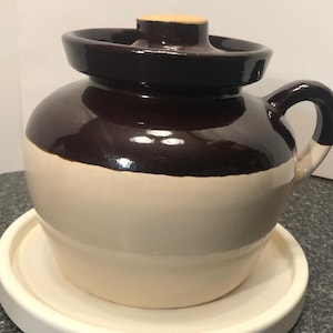 Puede incluir: Un tarro de cerámica esmaltado marrón y blanco con tapa. El tarro tiene un cuerpo redondeado y un asa en el lateral.