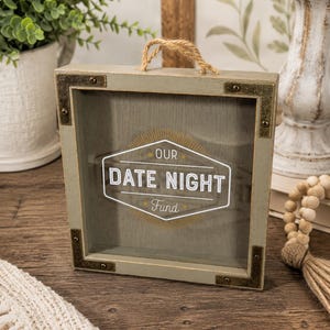 May include: A gray wooden shadow box with a clear front, metal corner accents, and a rope handle. The box features the text "OUR DATE NIGHT Fund" in a white and gold design, perfect for saving money for date nights.
