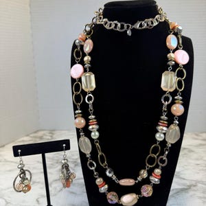 Könnte beinhalten: Ein mehrreihiges Halsketten-Set mit passenden Ohrhängern, verziert mit verschiedenen Perlen in Pfirsich-, Rosa- und klaren Glastönen, durchsetzt mit silber- und bronzefarbenen Metallringen und Kettengliedern für eine mehrschichtige Optik.