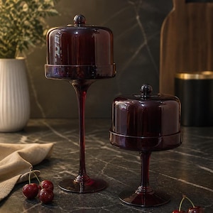 May include: Two burgundy glass cake stands with matching dome lids. One stand is tall, the other shorter. Fresh cherries sit nearby on a dark countertop. A white vase with greenery and a wooden cutting board are in the background.