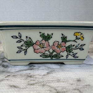 Peut inclure: Un pot de fleurs rectangulaire en céramique avec une base de couleur crème et des bords évasés. L'extérieur présente un motif floral peint à la main avec deux grandes fleurs roses, des feuilles vertes et une fleur jaune, souligné de lignes bleues.