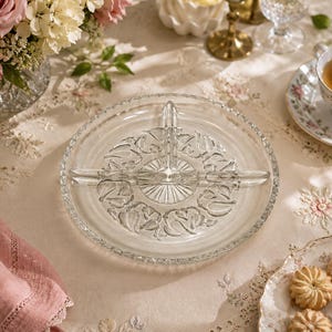 Peut inclure: Un plat de service en verre transparent divisé en trois sections, orné d'un motif floral. Le plat rond présente un bord décoratif. L'image comprend également une tasse à thé, des biscuits et des fleurs, évoquant une ambiance de goûter.