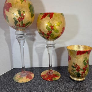 May include: Three decorative glass candle holders. Two are tall, with clear stems and round bases decorated with red poinsettias. The globes are gold with red poinsettias, green leaves, and red berries. The third is a short, round candle holder with the same design.