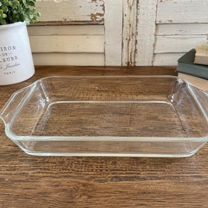 Puede incluir: Una fuente de horno rectangular de vidrio transparente con asas. La fuente está vacía y se encuentra sobre una superficie de madera. Una maceta de cerámica blanca con el texto "MAISON DE FLEURS Le Jardin PARIS" está en el fondo.