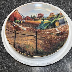 May include: Decorative plate featuring a rural farm scene with a red barn, tractors, and pheasants in flight. The plate has a gold rim and is set in a white protective holder. The artwork depicts a harvest scene with autumn colors.