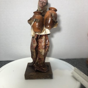 May include: A handmade paper mache figurine of a woman carrying two brown clay pots. The woman is wearing a white head covering and a brown dress. The figurine is standing on a wooden base.