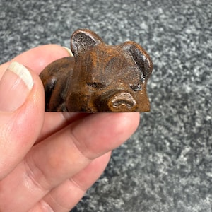 May include: A small, brown, sleeping pig figurine. The pig is lying down with its eyes closed and ears perked up. The figurine is made of a dark brown material and has a textured surface.