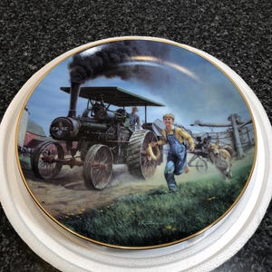 Puede incluir: Plato decorativo con una escena rural con un tractor de vapor antiguo, un niño corriendo y un perro. El plato tiene un borde dorado y está sobre un cielo azul con humo oscuro. El tractor tiene el texto "CASE".